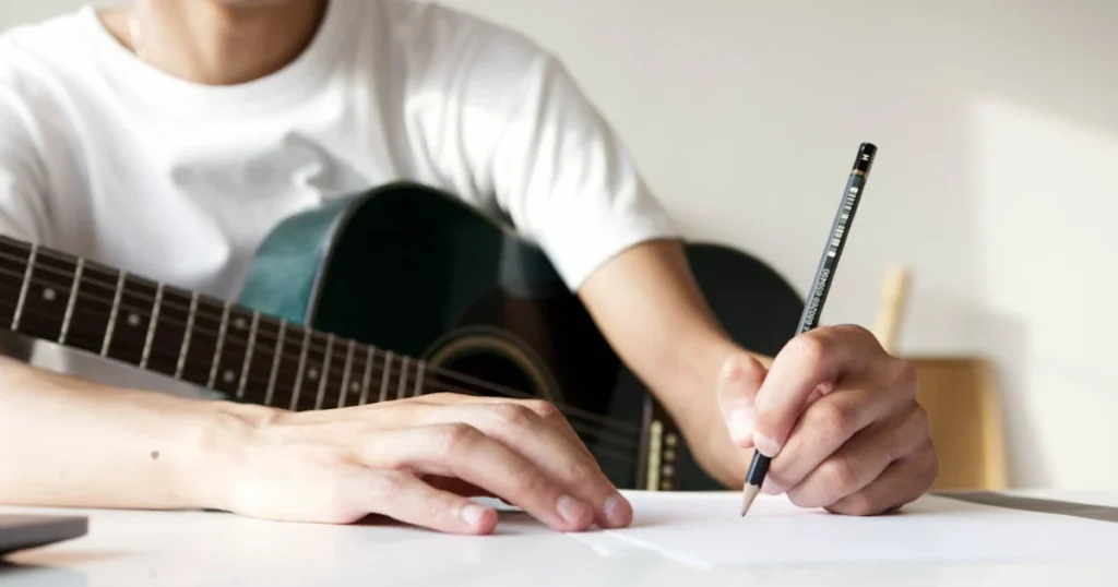Imagem de uma pessoa com um violão escrevendo com um lápis preto em uma folha de papel, talvez escrevendo música